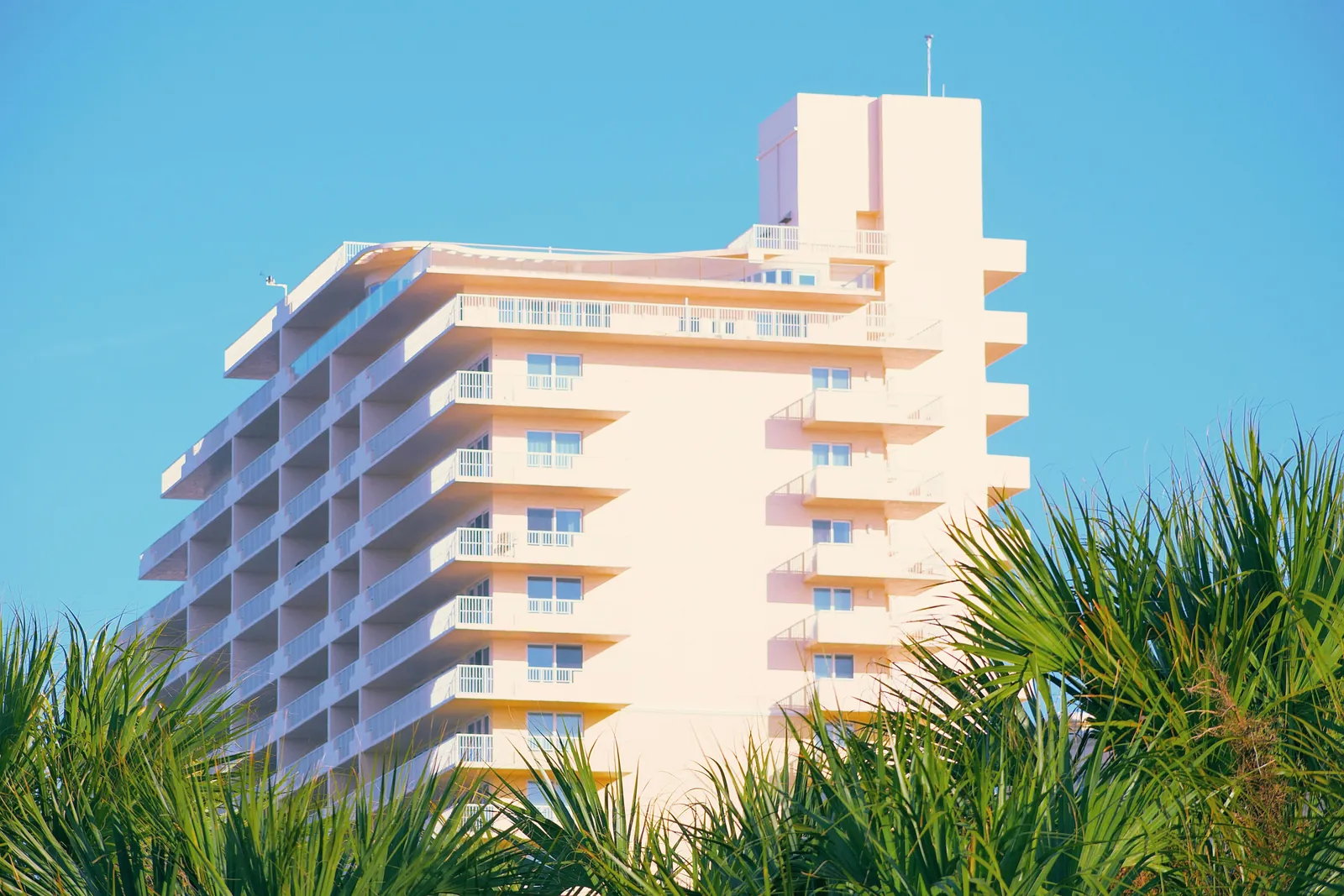A pale peach hotel tower with stacked balconies behind palm fronds under a blue sky