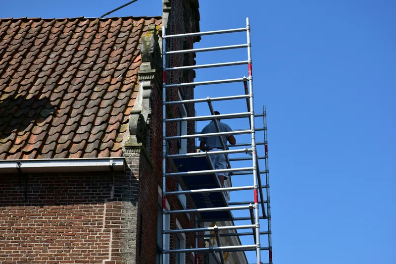 A tradesman on aluminium scaffolding beside a brick building with a tiled roof