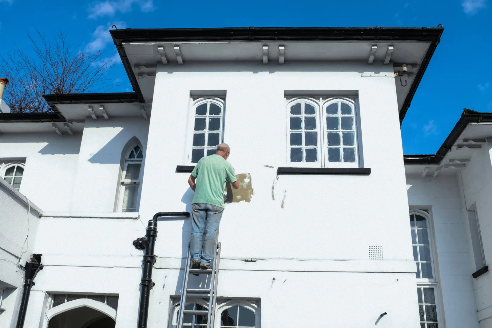 A painter on a ladder working on the white facade of a two-storey building under a blue sky