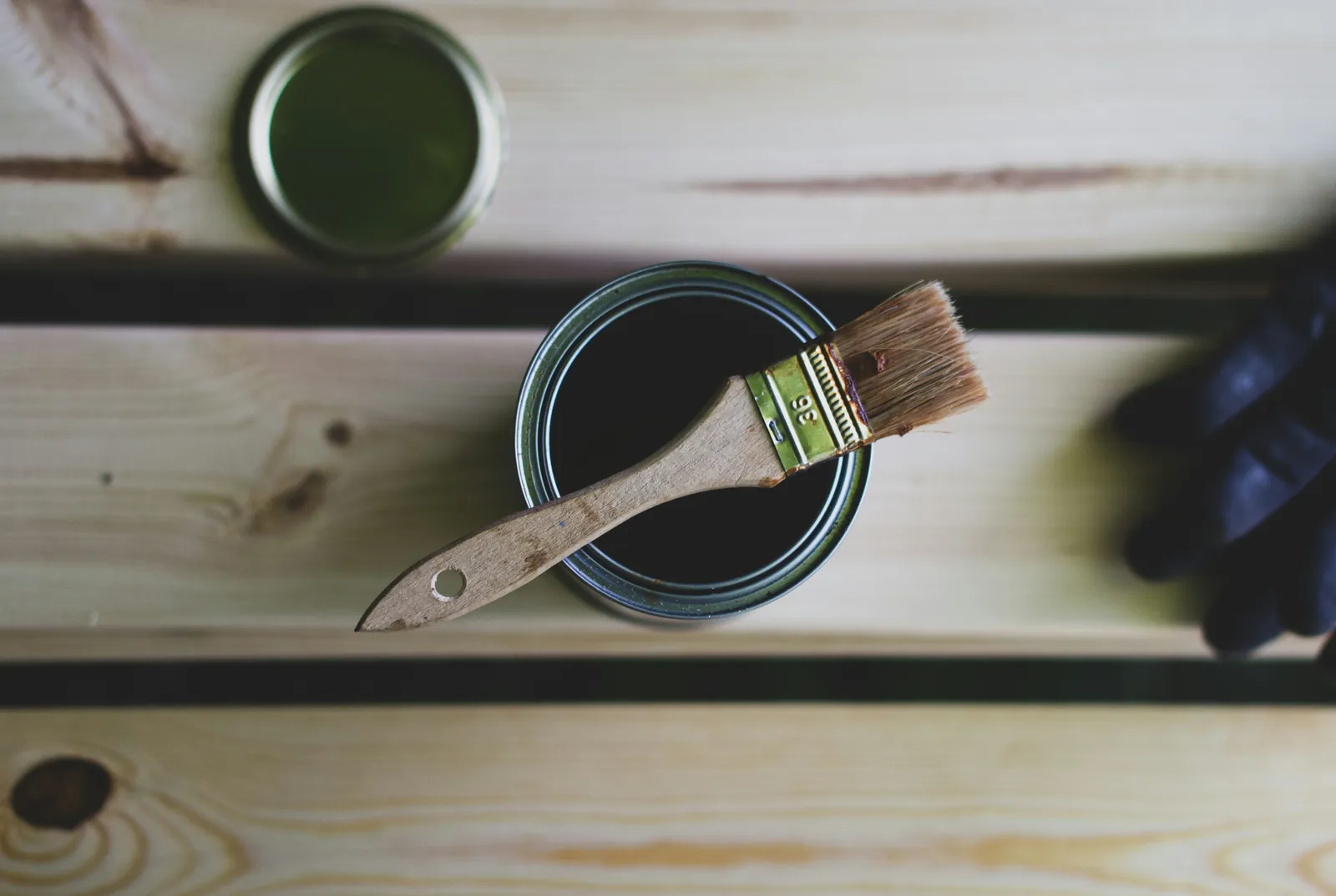A paintbrush resting on an open tin of wood stain on light wooden planks